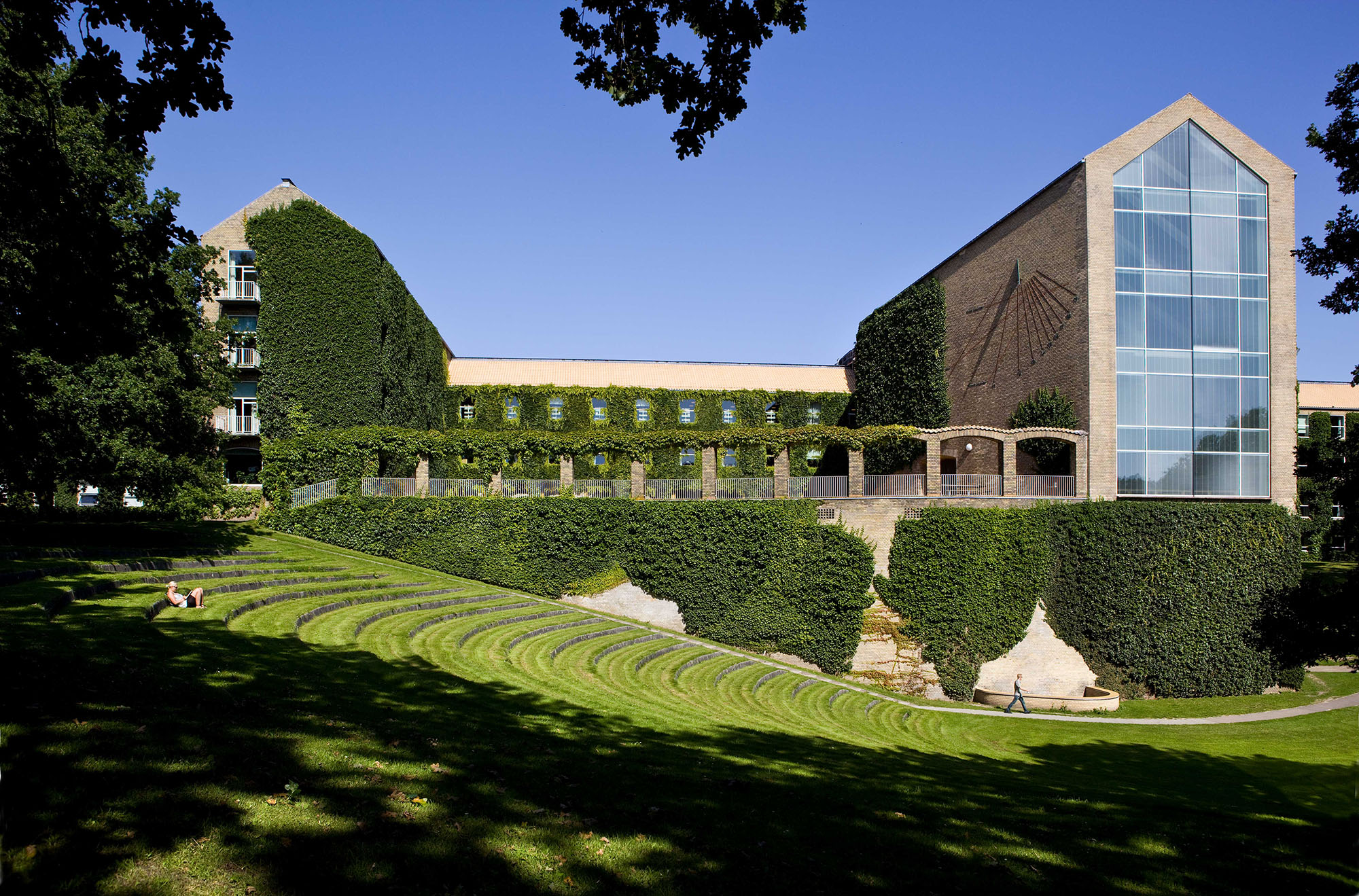 A landscape photograph of a brick Aarhus University building covered in ivy and grassy landscape stairs in the foreground. Trees frame the left and right sides.