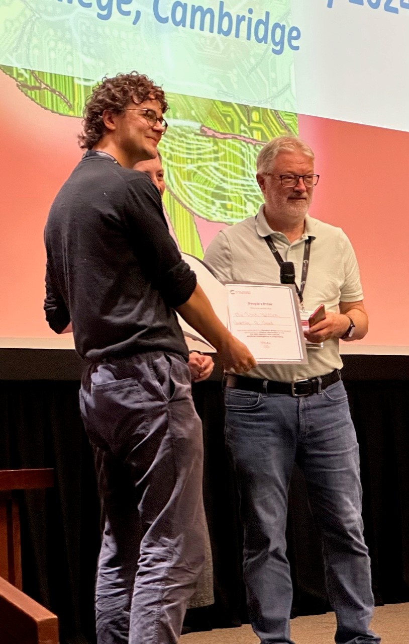 Three white men stood on a stage. One, closest to the camera, has curly hair and wears glasses, is holding a certificate which is being presented to him by the other two men who appear to be older due to their grey and white hair.